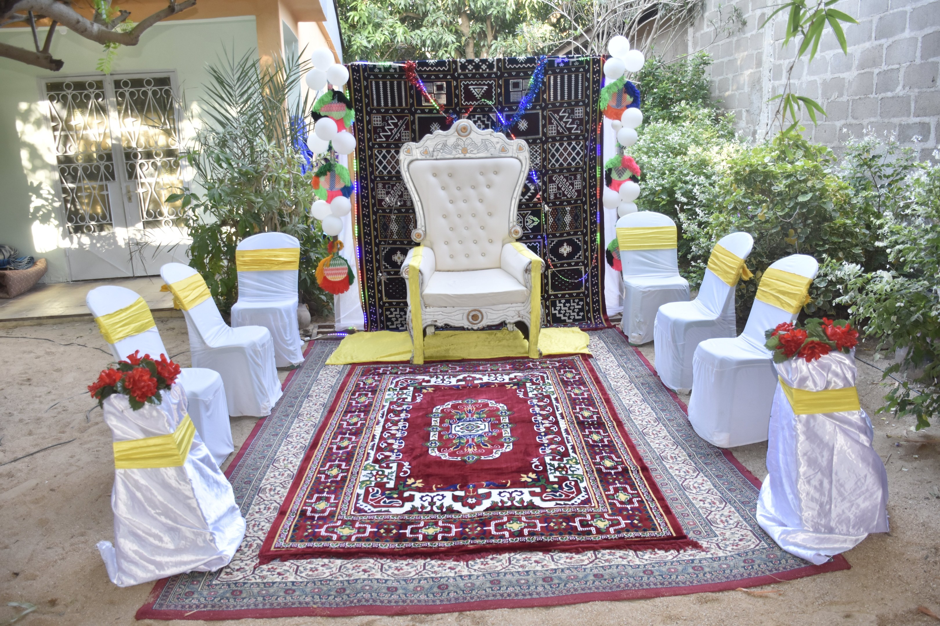 Eine Gartenhochzeitsaufstellung mit weißen Stühlen, einem roten Teppichläufer, Blumenbouquets, dekorativen Elementen, Luftballons, Pflanzen, Bäumen, einem Gebäude mit Fenstern und einer Tür und einer Wand auf der rechten Seite.