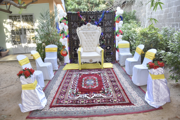 Eine Gartenhochzeitsaufstellung mit weißen Stühlen, einem roten Teppichläufer, Blumenbouquets, dekorativen Elementen, Luftballons, Pflanzen, Bäumen, einem Gebäude mit Fenstern und einer Tür und einer Wand auf der rechten Seite.