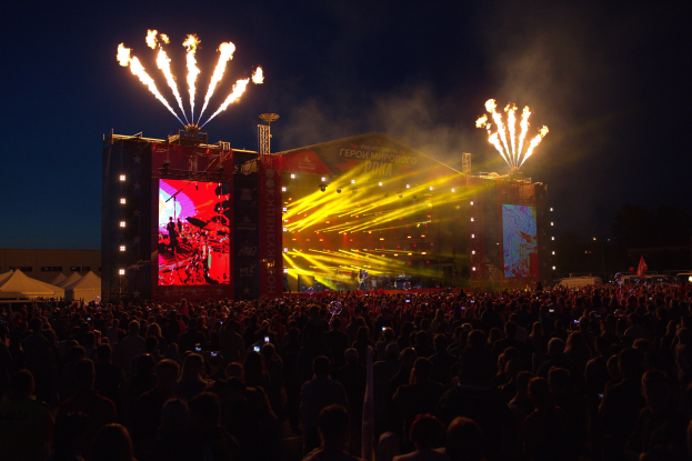 Eine große Menge bei einem Musikfestival schaut auf einer hell erleuchteten Bühne mit Bannern, umgeben von Zelten und Feuerwerk am Nachthimmel.