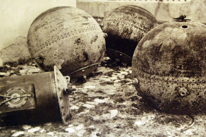 Drei große Metallbomben stehen vor einer Wand mit einem Fenster im Hintergrund in einem Schwarz-Weiß-Foto.