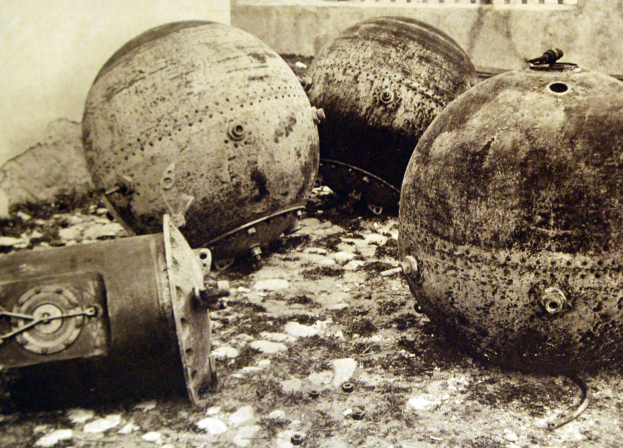 Drei große Metallbomben stehen vor einer Wand mit einem Fenster im Hintergrund in einem Schwarz-Weiß-Foto.