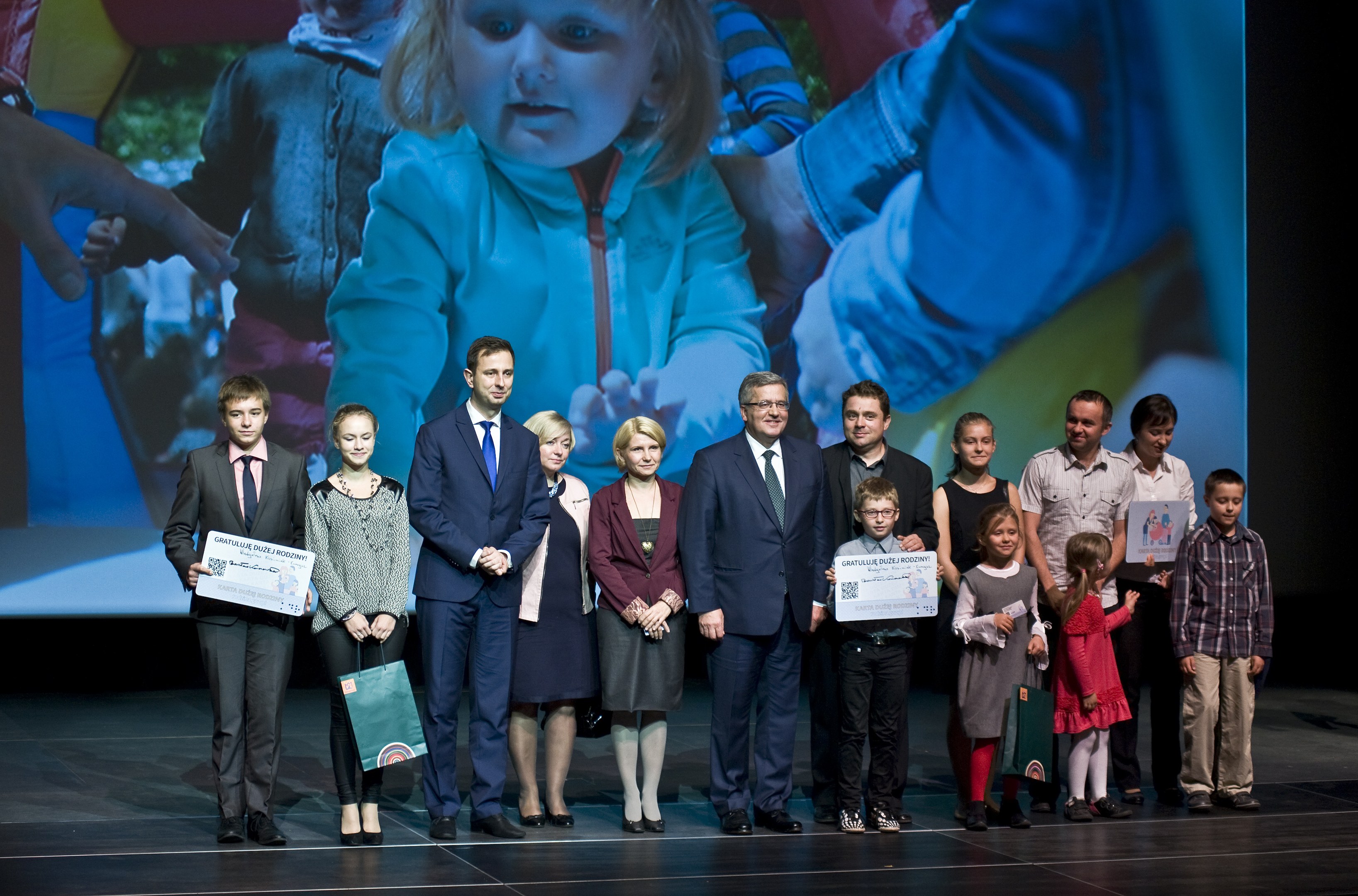 Gruppe von Menschen auf einer Bühne mit Taschen und Urkunden während eines Wohltätigkeitsereignisses, im Hintergrund ein Bildschirm mit einem Bild eines Kindes und anderen.