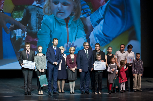 Gruppe von Menschen auf einer Bühne mit Taschen und Urkunden während eines Wohltätigkeitsereignisses, im Hintergrund ein Bildschirm mit einem Bild eines Kindes und anderen.