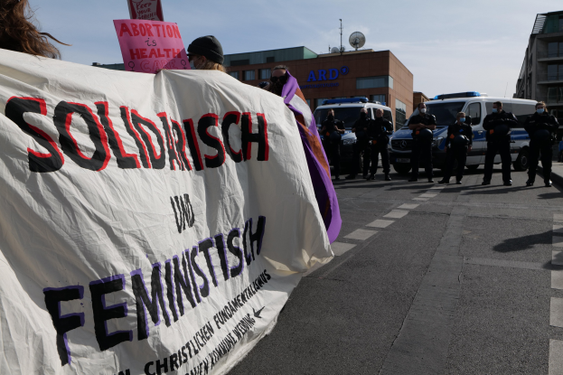 Eine Gruppe von Menschen marschiert die Straße entlang und hölt eine "Solidarität und Feminismus"-Fahne, mit geparkten Fahrzeugen, Gebäuden, einer Schüsselantenne und einem klaren blauen Himmel im Hintergrund.