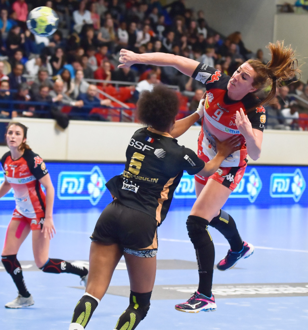 Eine Gruppe von Frauen spielt Handball auf einem Feld mit einem Ball in der Luft, in einem Stadion mit Zuschauern im Hintergrund.