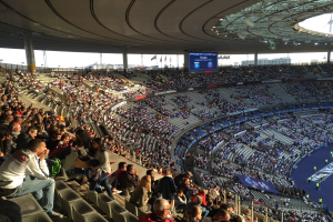 Große Menschenmenge in der Allianz Arena bei einem Fußballspiel sitzend, mit einer Bühne auf der rechten Seite, Fahnen, Stangen, einem Bildschirm und sichtbarem Himmel im Hintergrund.