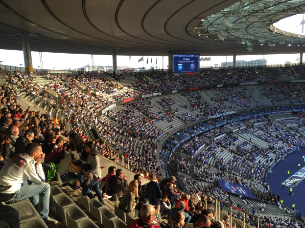 Große Menschenmenge in der Allianz Arena bei einem Fußballspiel sitzend, mit einer Bühne auf der rechten Seite, Fahnen, Stangen, einem Bildschirm und sichtbarem Himmel im Hintergrund.