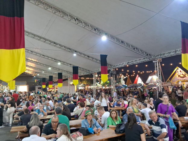 Große Gruppe von Menschen sitzt an langen Tischen unter einem Zelt auf dem Oktoberfest in München, Deutschland, mit Taschen und Gegenständen auf den Tischen und festlicher Dekoration im Hintergrund.
