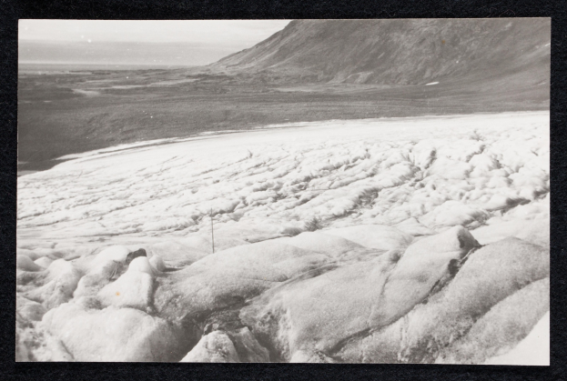 Schwarz-weiß gerahmtes Foto eines großen Gletschers mit schneebedeckter Landschaft und einem Hügel im Hintergrund.