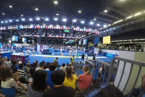 Große Zuschauermenge sitzend und stehend in einer Halle mit Bildschirmen, Fahnen und Deckenleuchten im Hintergrund.