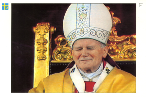 Ein Mann in priesterlicher Kleidung und einem weißen Hut sitzt auf einem Stuhl vor einem dunklen Hintergrund und strahlt eine königliche und würdige Ausstrahlung aus.