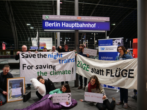 Eine Gruppe von Menschen sitzt auf dem Boden des Berliner Hauptbahnhofs und hält ein Banner mit der Aufschrift "Retten Sie die Nachtzüge zum Schutz des Klimas", umgeben von Kissen, einem Foto Rahmen und anderen Gegenständen, mit Brettern, Säulen, Lampen und Geländern im Hintergrund.