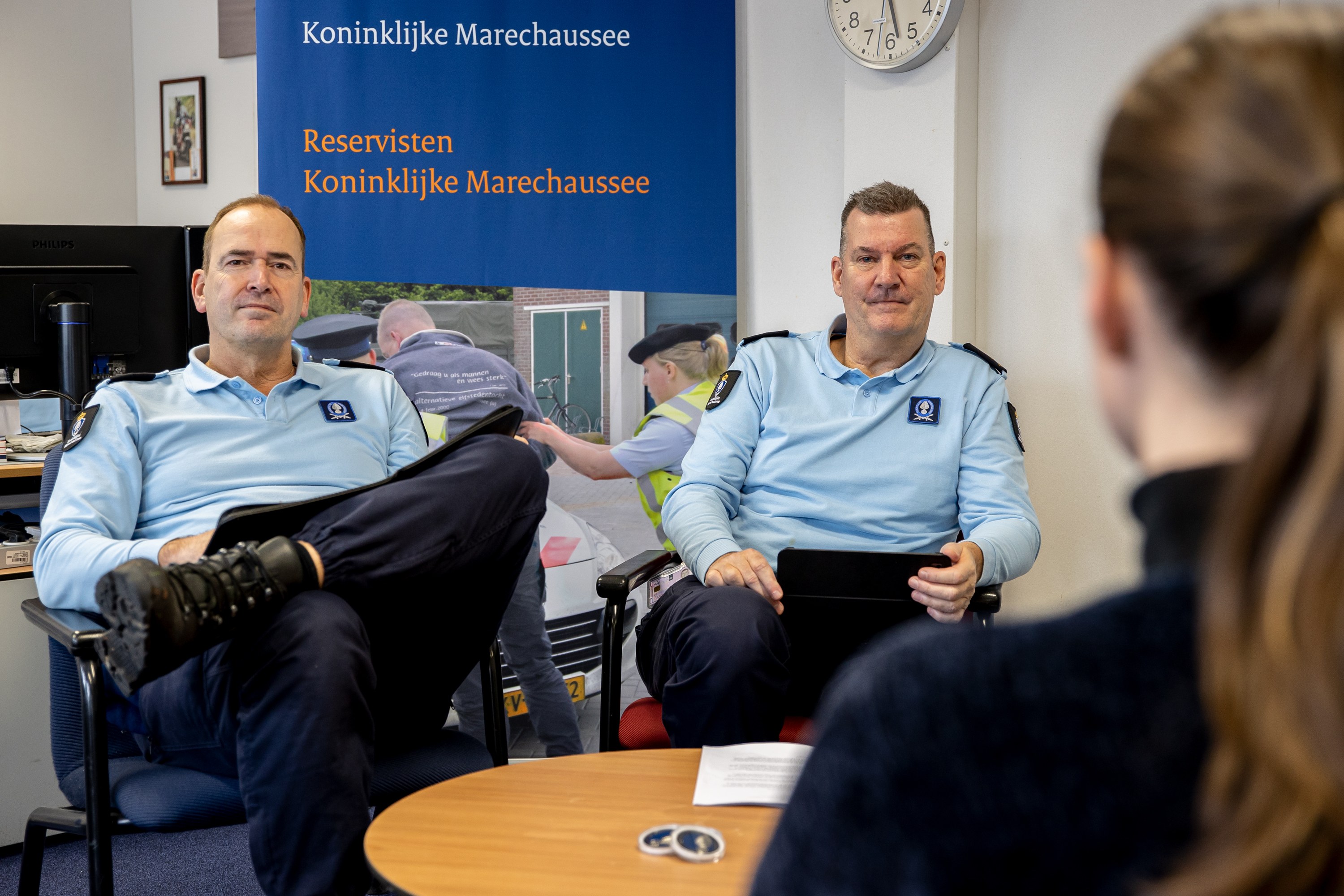 Zwei Polizeibeamte sitzen an einem Tisch mit einer Frau, Papiere und Gegenstände auf dem Tisch, eine Wandtafel und eine Uhr im Hintergrund, ein zweiter Tisch mit einem Monitor, Tastatur, Maus und Gegenständen links daneben und Fotorahmen an der Wand.