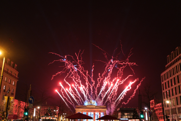 Eine belebte Straßenkreuzung in Berlin an Silvester, voller Menschen, Fahrzeuge und Gebäude, beleuchtet von Feuerwerk und Gebäudelichtern, die eine festliche Atmosphäre schaffen.