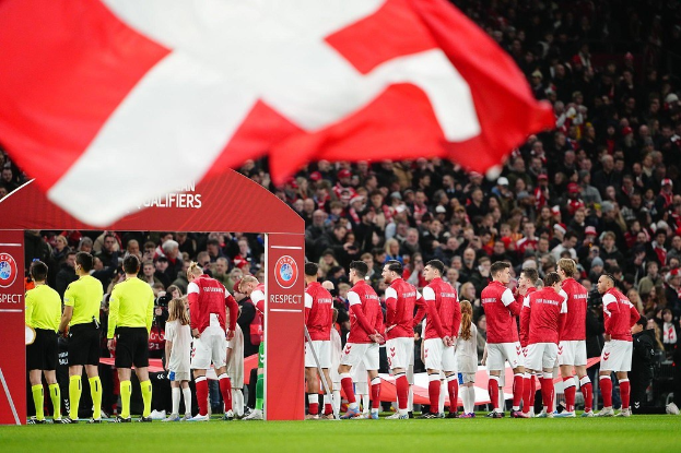 Gruppe von Menschen auf einem Fußballfeld mit einer roten und weißen Fahne im Vordergrund, einem Bogen mit der Aufschrift "Bayern Munich vs Bayern Munich Betting Tips & Preview" im Hintergrund und einer großen Stadionmenge.