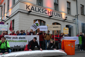 Eine Gruppe von Menschen mit Protestschildern und Transparenten vor einem Gebäude, mit zwei Personen im Vordergrund auf einem Gegenstand sitzend und einem Müllcontainer rechts, vor einem Hintergrund von Gebäuden mit Fenstern und Schildern in Deutschland.