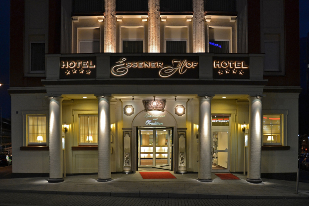 Das Essenerhof Hotel in Berlin, Deutschland, ein mehrstöckiges Gebäude mit Fenstern, Türen, Säulen und Lichtern, mit einem Namensschild sichtbar, und einer Straße mit Fahrzeugen rechts daneben, unter einem klaren Himmel.