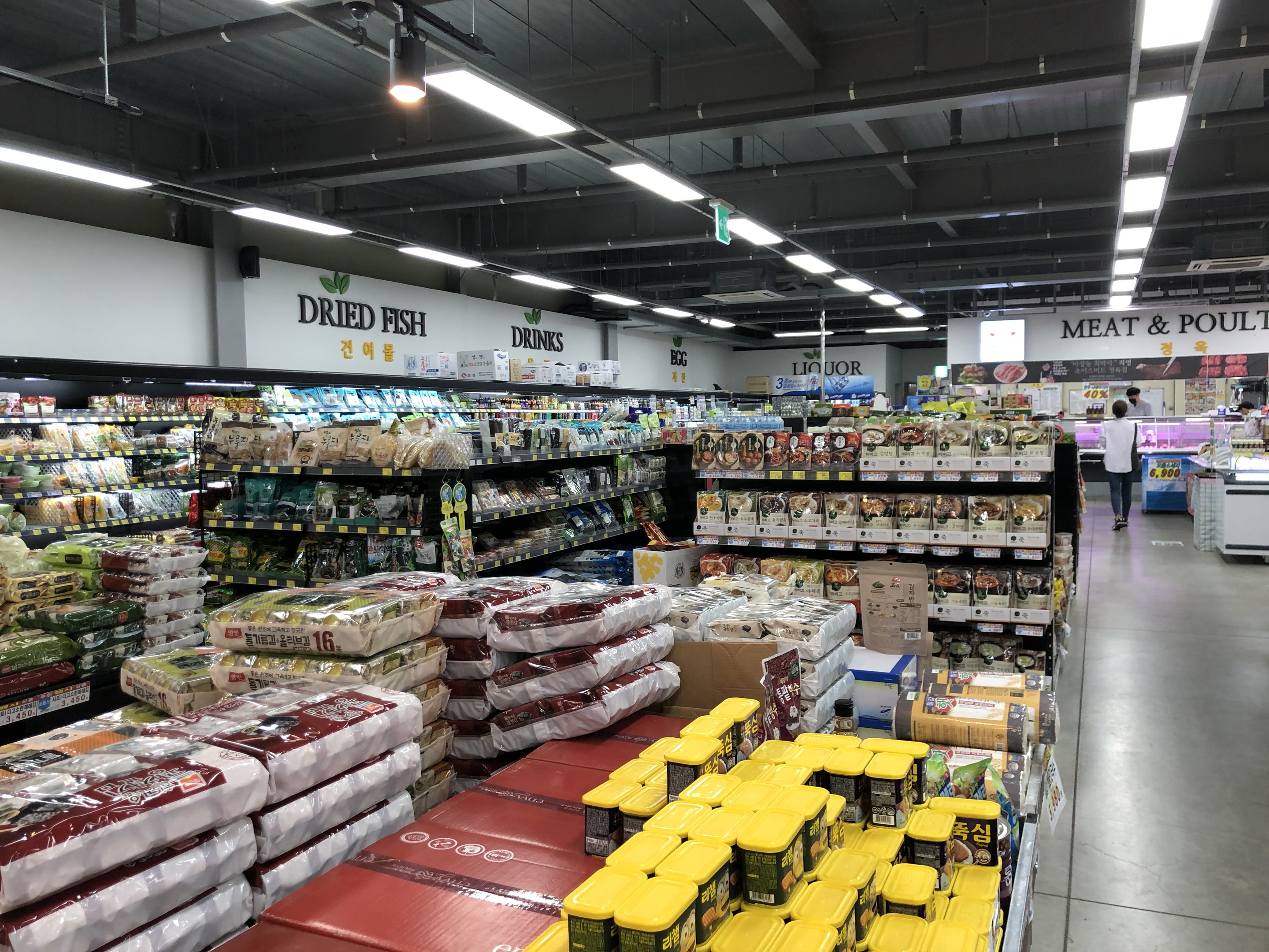 Ein Supermarktgang mit verpackten Lebensmitteln auf Regalen, mit Texttafeln, einer Person, einem Tisch und Deckenleuchten.