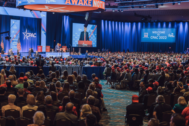 Eine große Menschenmenge sitzt auf Stühlen, die einer Bühne zugewandt sind, mit einigen Menschen darauf, blaue Vorhänge, ein Bildschirm und eine orangefarbene Tafel oben, die das CPAC 2020-Ereignis am 25. Oktober anzeigt.