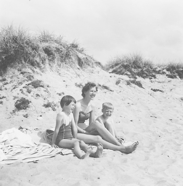 Schwarz-weißes Foto einer Frau in einem Bikini und zweier Kinder in Badebekleidung, die auf einer Decke am Strand sitzen, mit Sand, Gras und Himmel im Hintergrund.