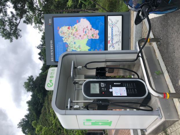 Eine Ladestation für Elektrofahrzeuge am Straßenrand mit einem Motorrad in der Nähe, ein digitales Anzeigetafel, die die Ladestation für E-Bikes anzeigt, und Bäume mit einem klaren blauen Himmel im Hintergrund.