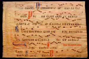 Ein illuminiertes Manuskript mit Musiknotation auf einem Blatt Papier vor einem dunklen Hintergrund, gefüllt mit Text und Musiksymbolen.