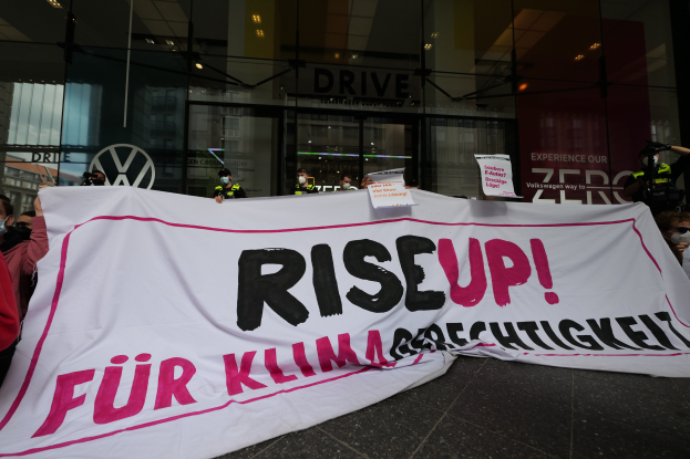Eine Gruppe von Menschen mit Masken, die ein 'Aufstehen'-Schild und Schilder vor einem Glasgebäude bei Nacht halten.