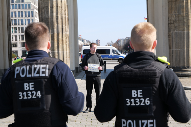 Drei Polizeibeamte stehen vor einem Gebäude, wobei ein Mann in der Mitte ein Papier hält, flankiert von Säulen, während Fahrzeuge, Menschen, Bäume, Gebäude und der Himmel im Hintergrund zu sehen sind.