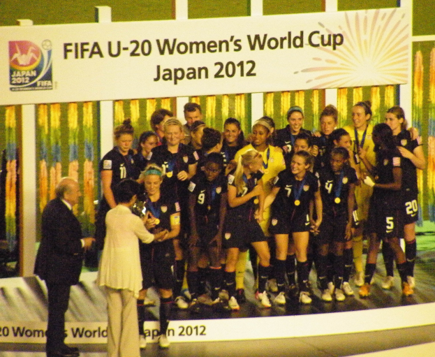 Gruppe von Frauenfußballspielerinnen in schwarzen Uniformen, die einen Pokal auf einer Bühne halten, mit einer Tafel im Hintergrund.