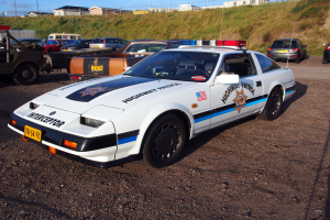 Ein Polizeiauto auf einem Parkplatz neben anderen Fahrzeugen, Gras, Pfosten, Drähte, Gebäude und einen klaren blauen Himmel.