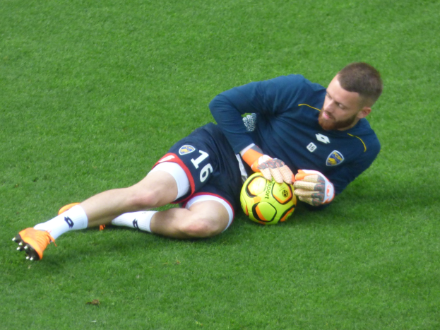 Ein Fußballspieler in einem blauen T-Shirt, Shorts, Socken und Schuhen liegt auf dem Boden und hält einen Ball.