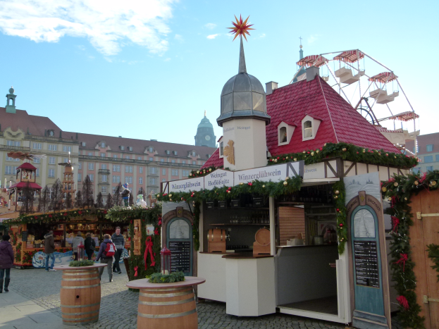 Ein lebendiger Weihnachtsmarkt in N├╝rnberg, Deutschland, mit Menschen um dekorierte St├Ąnde, Geb├Ąude, ein Riesenrad und einen bew├Âlktem Himmel sowie ein Schild auf der rechten Seite.
