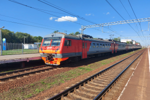Roter und oranger Zug fährt auf Schienen neben einem Bahnsteig mit einem Geländer, Bäumen, Masten, Drähten und einem klaren blauen Himmel im Hintergrund.