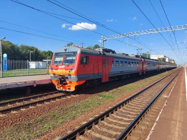 Roter und oranger Zug fährt auf Schienen neben einem Bahnsteig mit einem Geländer, Bäumen, Masten, Drähten und einem klaren blauen Himmel im Hintergrund.