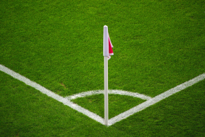 Ein Fussballfeld mit grünem Rasen, weißer Begrenzungslinien und einem Elfmeterpunkt, der durch einen weißen Pfosten mit einer rot-weißen Fahne markiert ist.