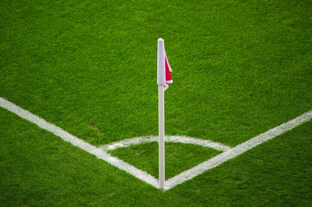 Ein Fussballfeld mit grünem Rasen, weißer Begrenzungslinien und einem Elfmeterpunkt, der durch einen weißen Pfosten mit einer rot-weißen Fahne markiert ist.