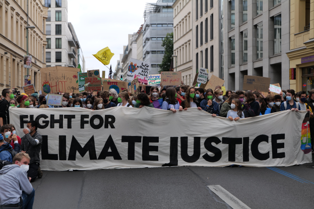 Eine Gruppe von Menschen marschiert mit einem "Kämpfe für Klimagerechtigkeit"-Schild und verschiedenen Plakaten die Straße entlang, trägt Masken und hat Gebäude auf beiden Seiten und einen klaren blauen Himmel im Hintergrund.