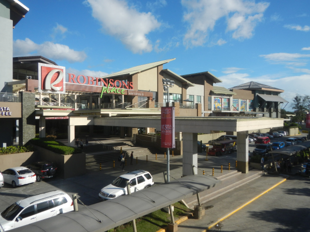 Ein belebter Einkaufszentrum mit verschiedenen Gebäuden, Geschäften, Fahrzeugen auf der Straße, Fußgängern auf dem Gehweg, Straßenlaternen, Informationsschildern, Grünflächen und einer bewölkten Himmel.