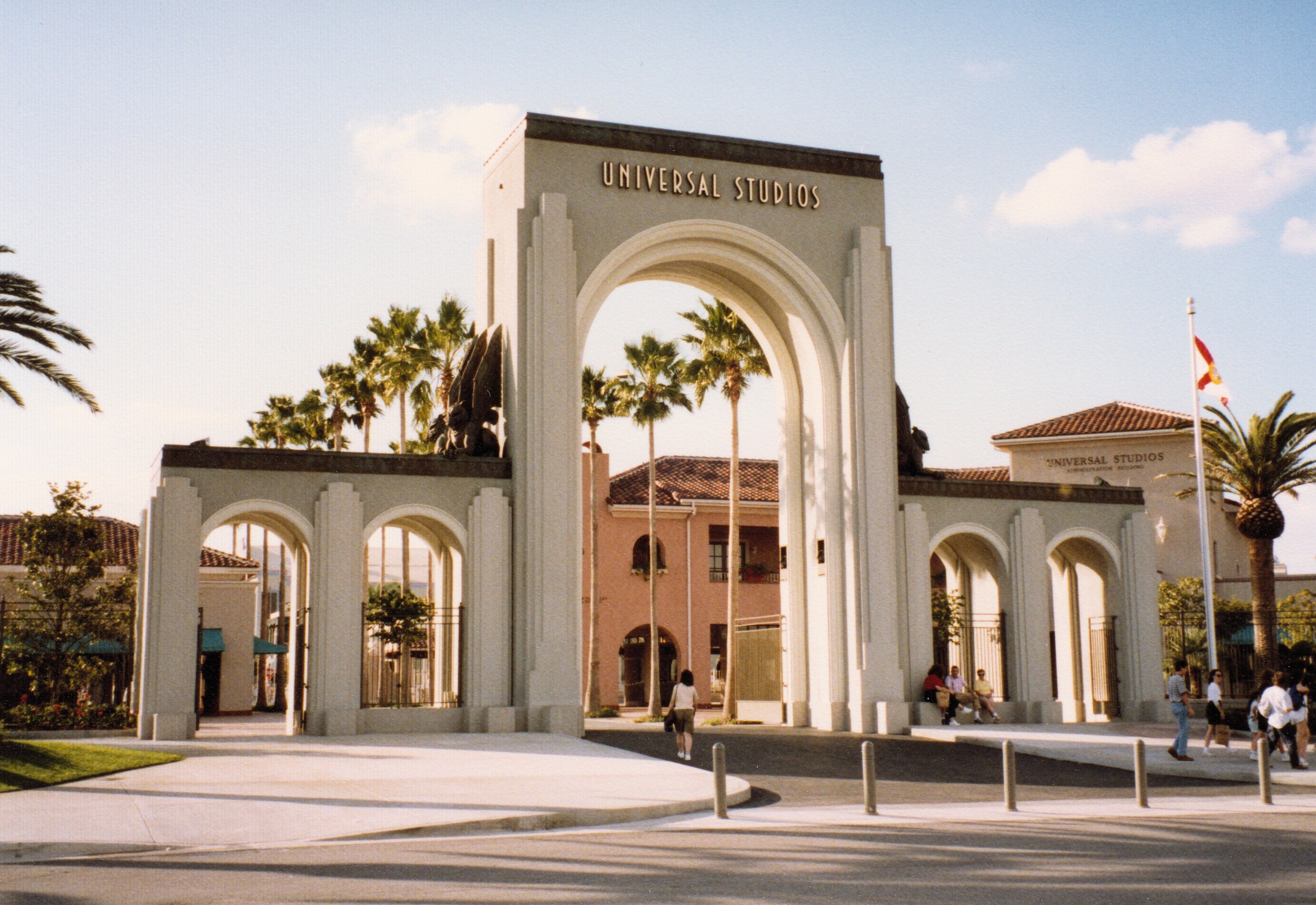 Eingang zu Universal Studios Hollywood mit einem Bogen, einer Statue, Besuchern, umgeben von Grün, Gebäuden, einem Fahnenmast und einem bewölkten Himmel.