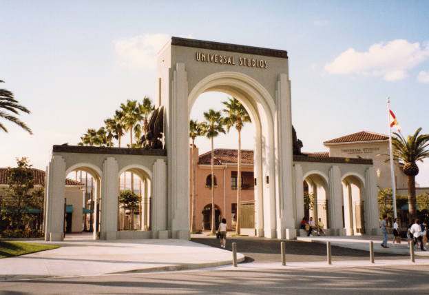 Eingang zu Universal Studios Hollywood mit einem Bogen, einer Statue, Besuchern, umgeben von Grün, Gebäuden, einem Fahnenmast und einem bewölkten Himmel.