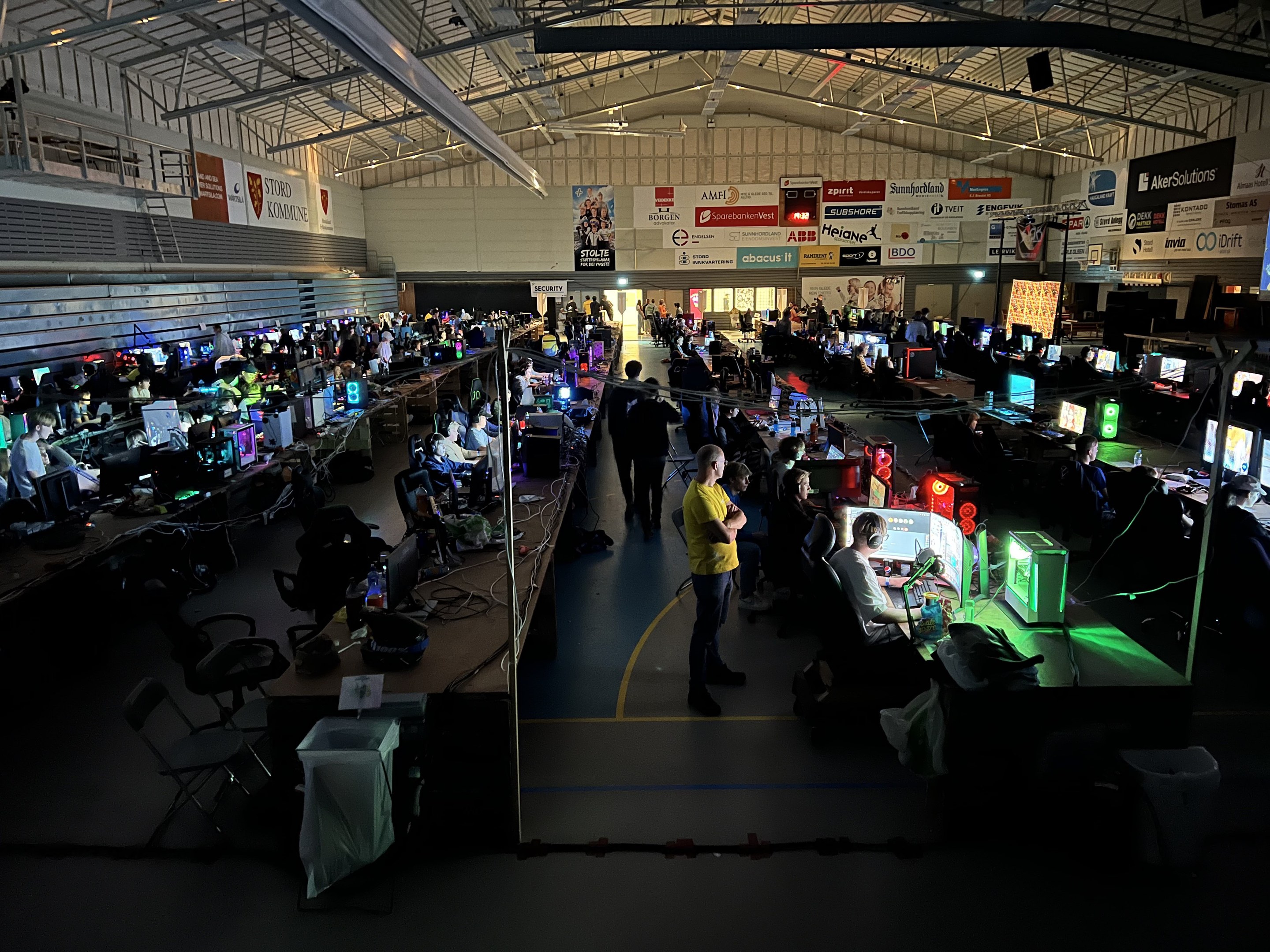 Ein großer Raum mit Menschen, die Videospiele spielen, sitzend und stehend, umgeben von Monitoren, Tastaturen, Mäusen und Kabeln auf Tischen, mit Tafeln im Hintergrund, die Text und Logos anzeigen.