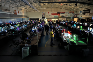 Ein großer Raum mit Menschen, die Videospiele spielen, sitzend und stehend, umgeben von Monitoren, Tastaturen, Mäusen und Kabeln auf Tischen, mit Tafeln im Hintergrund, die Text und Logos anzeigen.