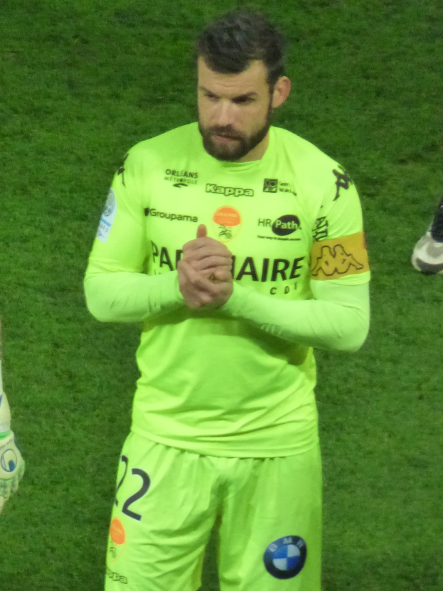 Torwart in gelbem Trikot klatscht auf dem Fußballfeld, trägt grünes T-Shirt und Shorts, mit Teil eines anderen Beins auf der linken Seite.