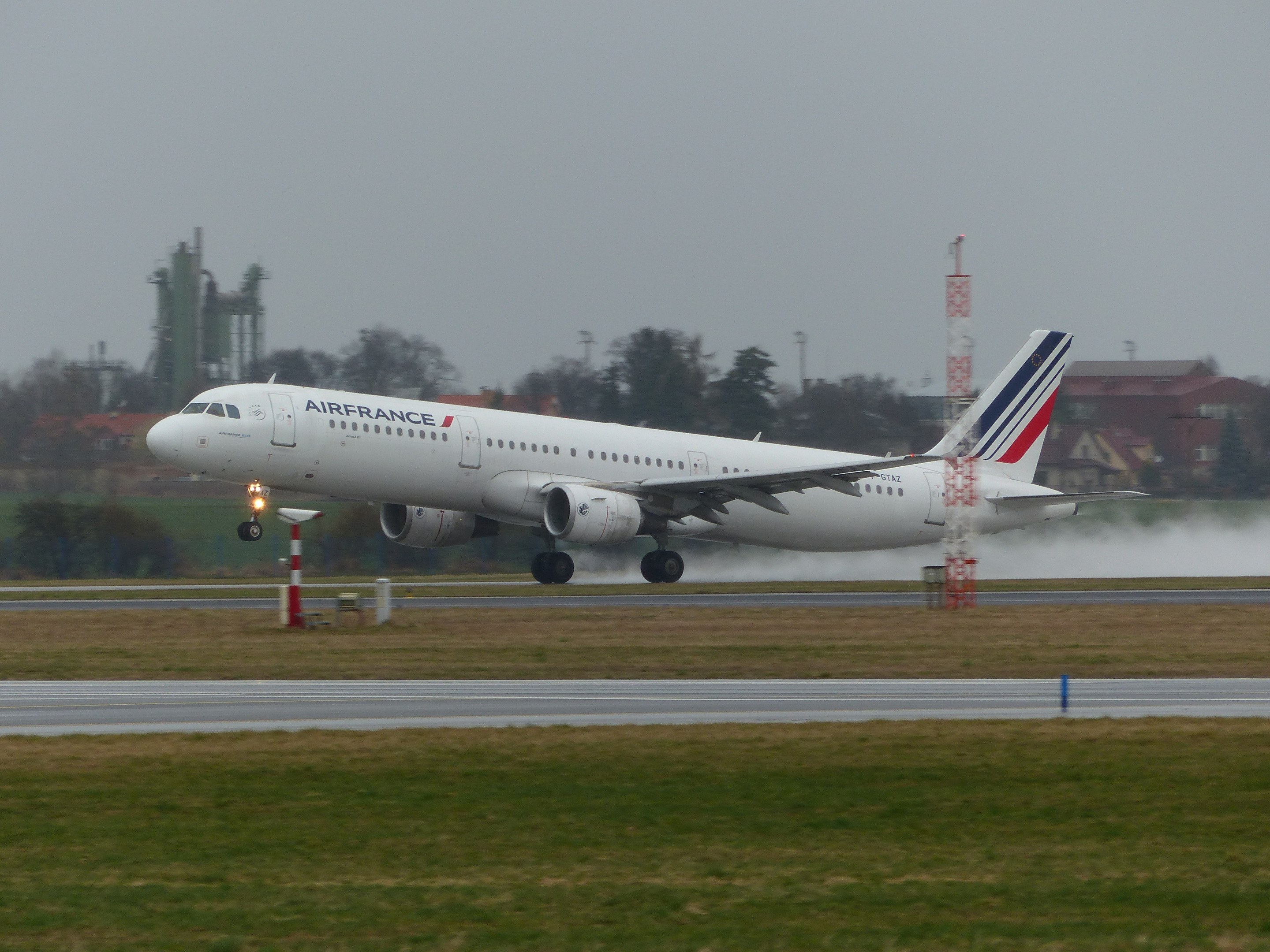 Air France Airbus A320-200 beim Starten vom Flughafen Paris Charles de Gaulle, umgeben von grünem Gras und Bäumen, mit Gebäuden und Türmen im Hintergrund unter einem klaren blauen Himmel.