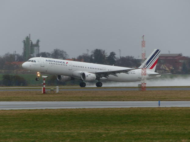 Air France Airbus A320-200 beim Starten vom Flughafen Paris Charles de Gaulle, umgeben von grünem Gras und Bäumen, mit Gebäuden und Türmen im Hintergrund unter einem klaren blauen Himmel.