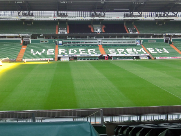 Ein großes Stadion mit einem grünen Feld, umgeben von Stühlen, Werbetafeln, Lampen, Stangen und einem drapierten Stuhl im Vordergrund.
