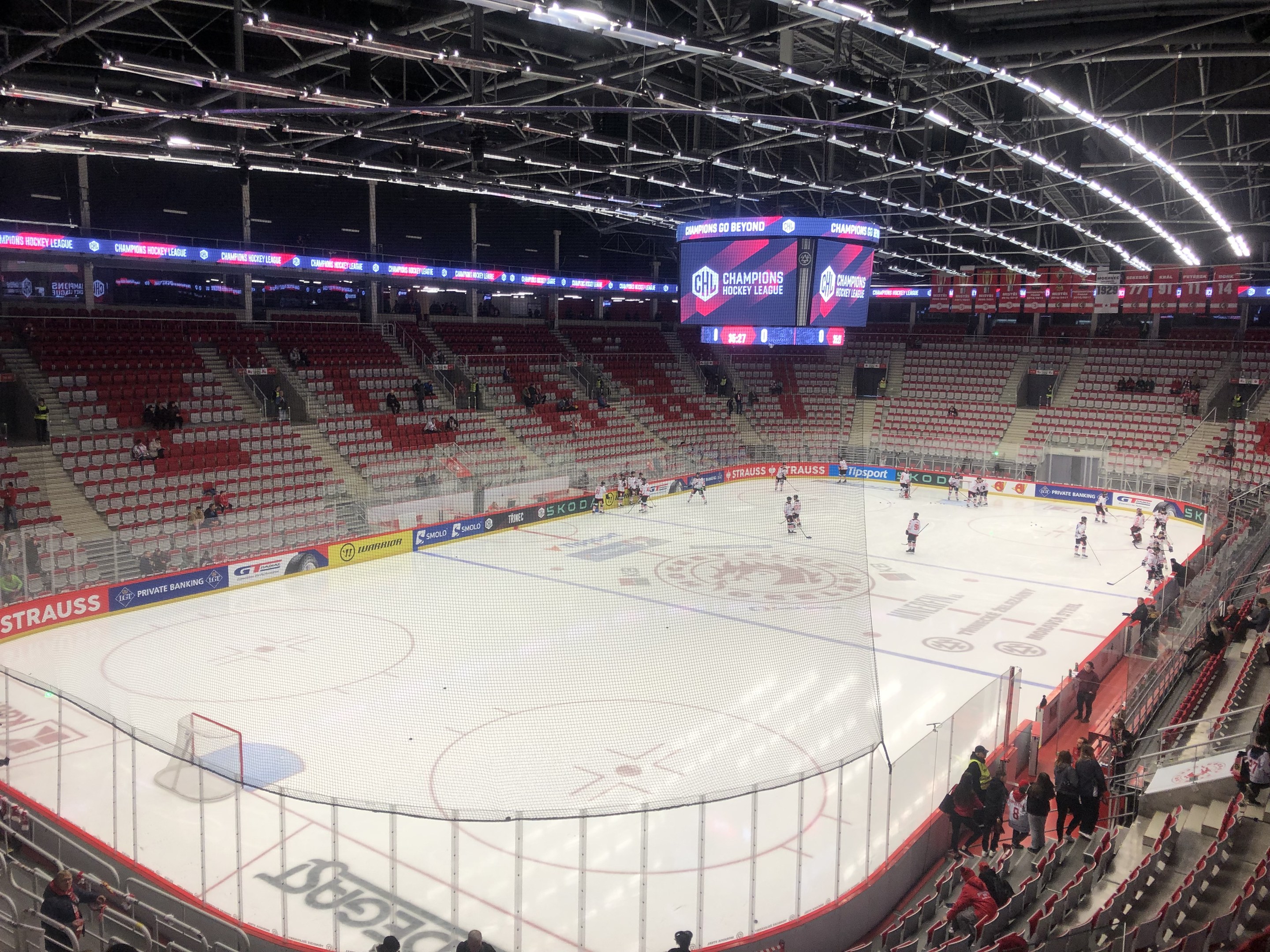 Eishockeyspiel in einer großen Arena mit Zuschauern, eine Eisfläche umgeben von einem Zaun, ein Display, das "St. Louis Blues vs Detroit Red Wings" anzeigt, Metallrahmen, Deckenlampen und ein Dach.