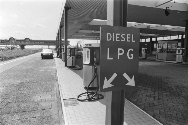 Schwarzes und weißes Foto einer Tankstelle mit einem "Diesel LPG"-Schild, Zapfsäulen und einem geparkten Auto vor einer Brücke, Bäumen, Gebäuden und einem bewölkten Himmel.