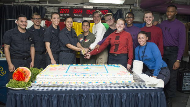 Eine Gruppe strahlender Menschen um einen Tisch mit einem Kuchen, Lebensmitteln und einer Wassermelone versammelt, die in einem Raum mit Vorhängen, einer digitalen Anzeigetafel, Lampen, Rohren und einer Wand feiern.