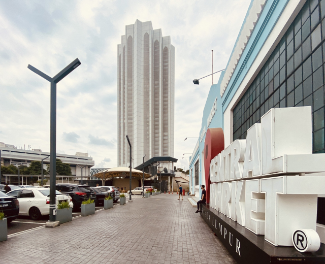 Ein belebter zentraler Markt in Kuala Lumpur, Malaysia, mit hohen Gebäuden, Straßenmasten, Laternen, Fahrzeugen, Fußgängern, Schildern, Bäumen und einem bewölkten Himmel.